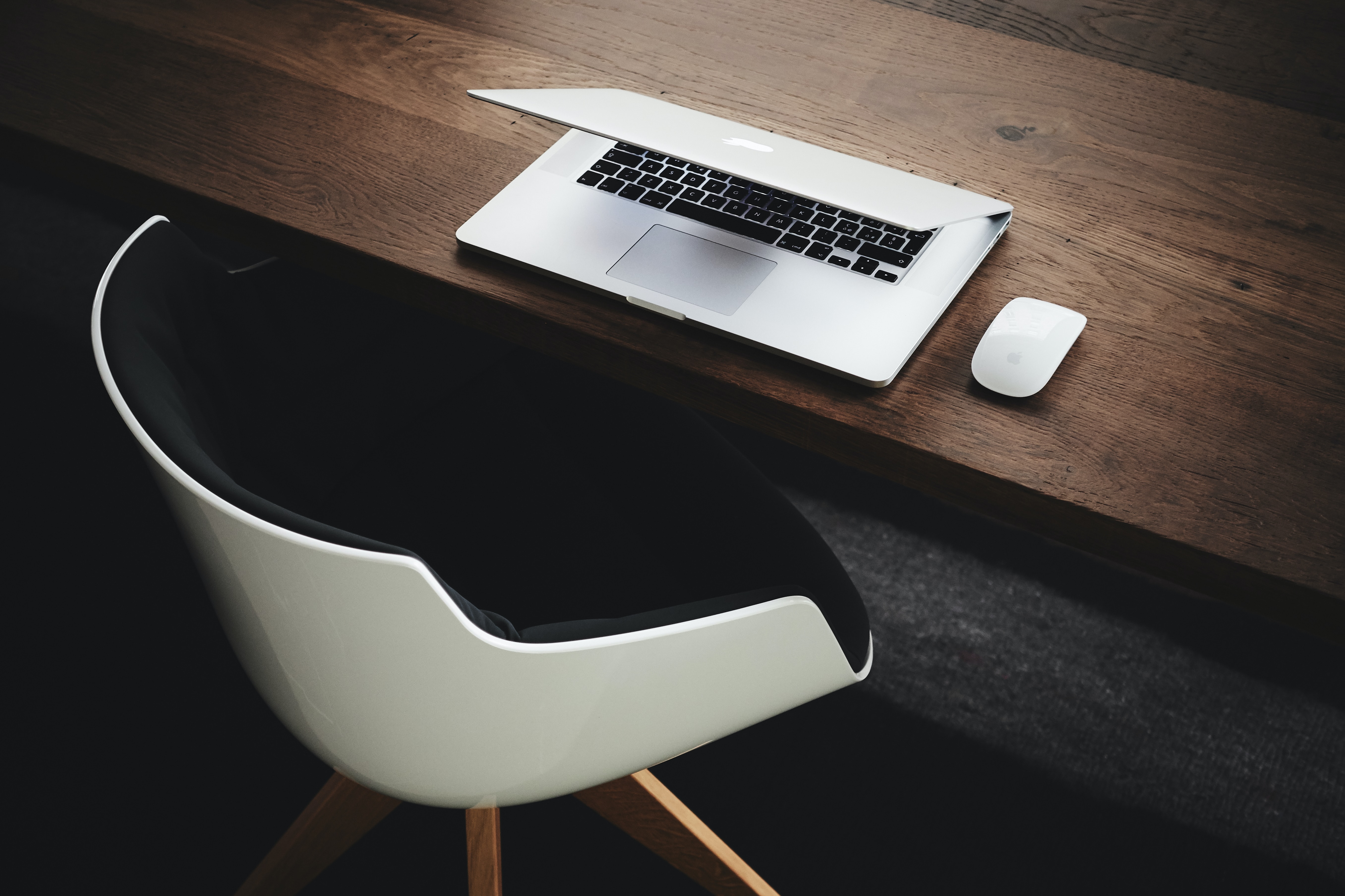 Silver Macbook closed midway on a wooden table having a modern white chair close by, photo credit to Luca Bravo