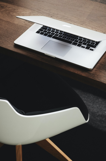 Silver Macbook closed midway on a wooden table having a modern white chair close by, photo credit to Luca Bravo